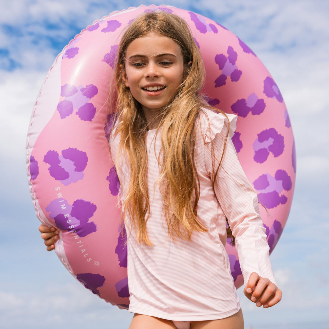 Young girl holding a pink and purple doughnut inflatable ring against a blue sky.