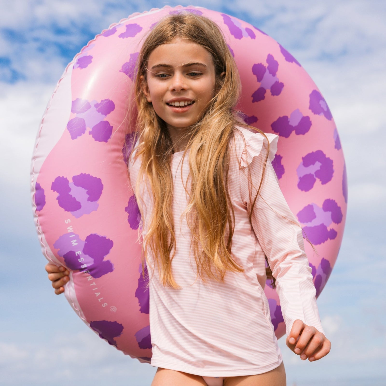 Young girl holding a pink and purple doughnut inflatable ring against a blue sky.
